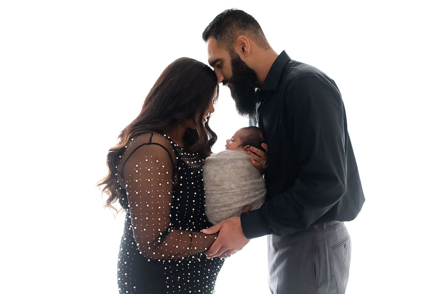 A couple in elegant attire share an intimate moment with their swaddled newborn.