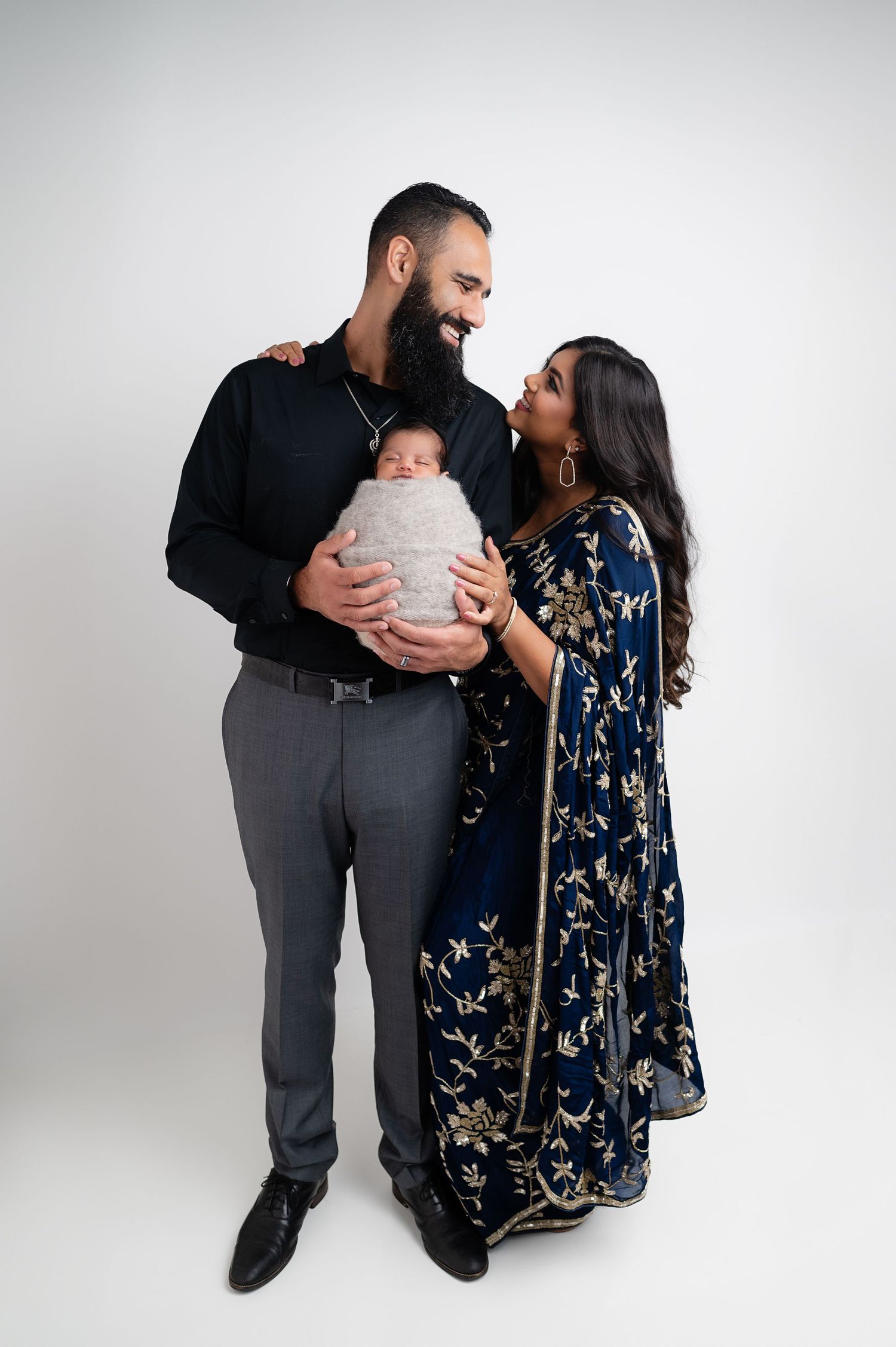 Family portrait with couple in formal attire holding their swaddled baby against white background.