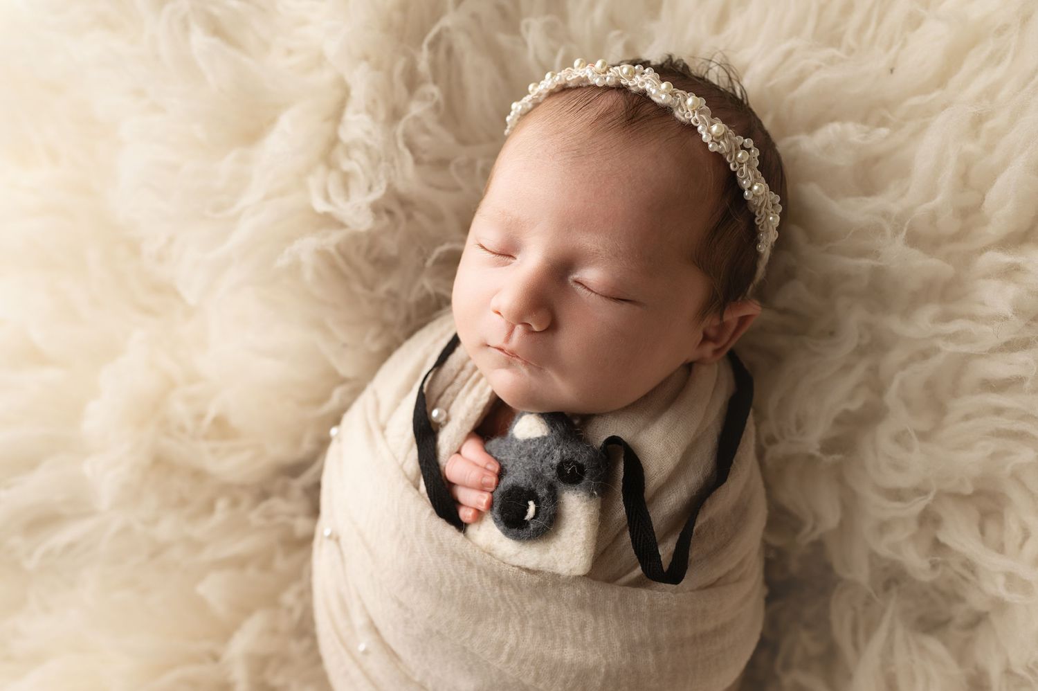 Newborn baby wrapped in white swaddle wearing bow headband and black bow tie lying on fluffy cream rug.