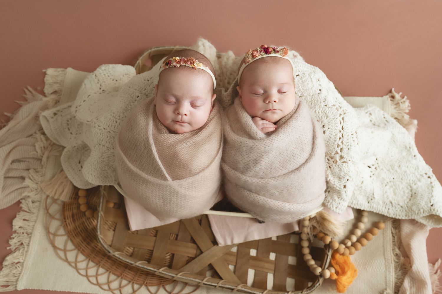 Twin newborns swaddled in beige wraps sleeping side by side in rustic basket.