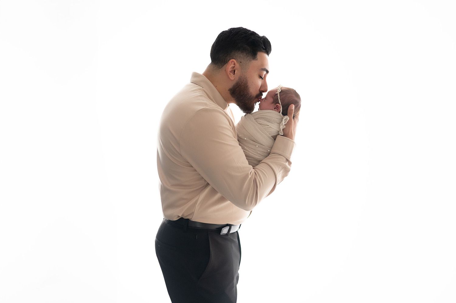 Parent in beige shirt gently holding and kissing newborn against white backdrop.