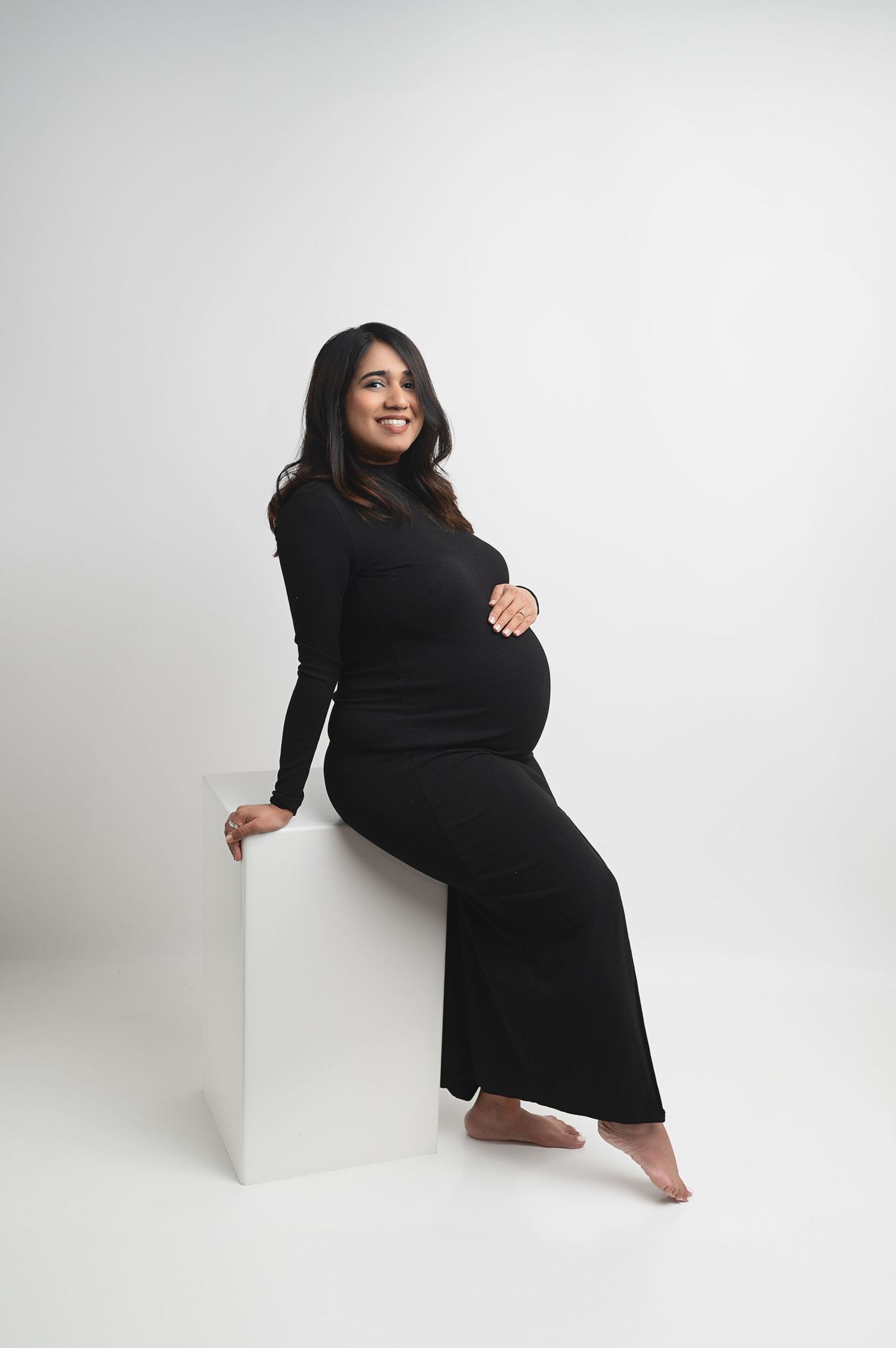 Expectant mother in black dress smiling while sitting on white cube during maternity photoshoot.