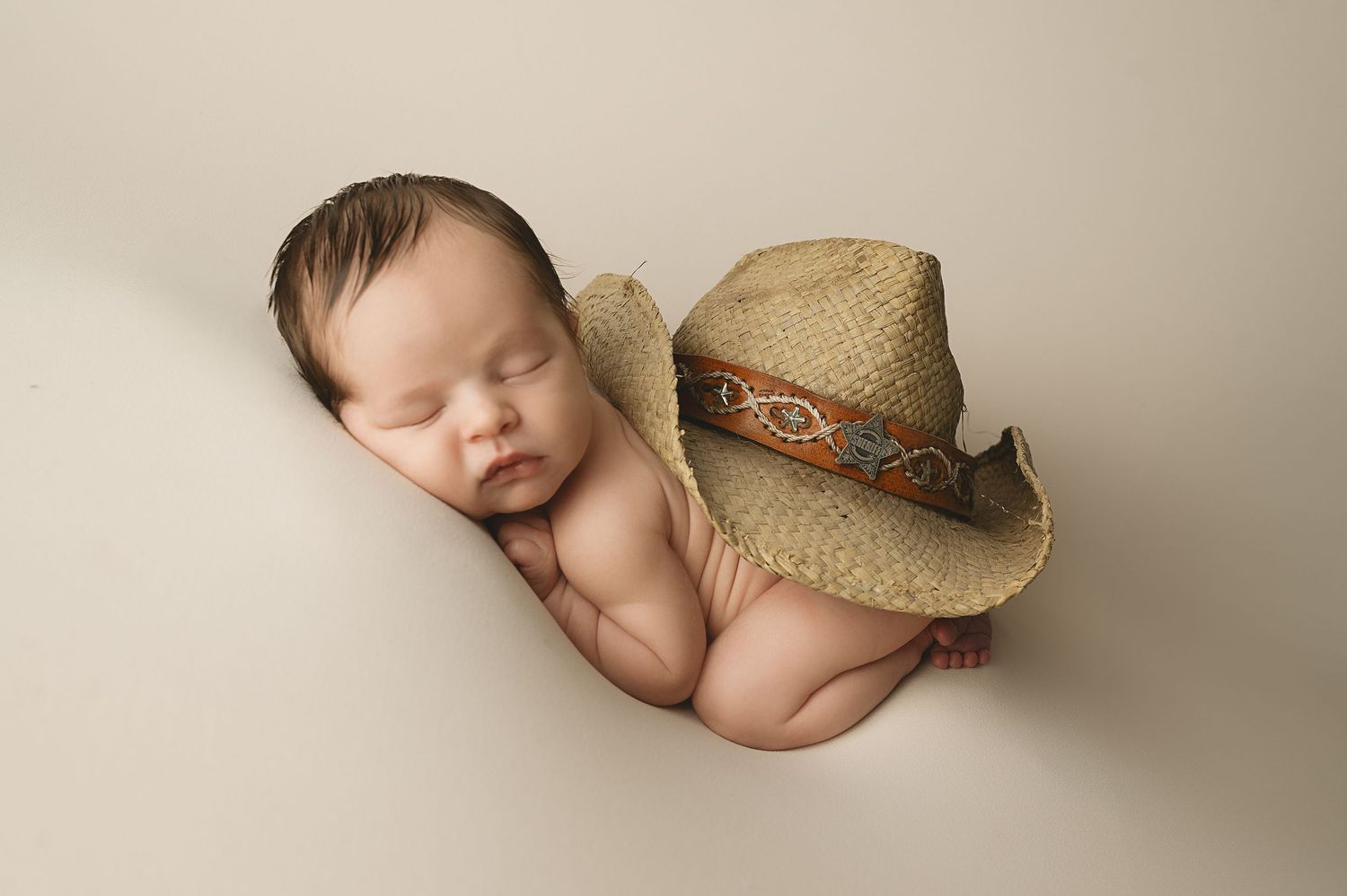 Sleeping newborn rests peacefully on cream backdrop with light brown straw cowboy hat.