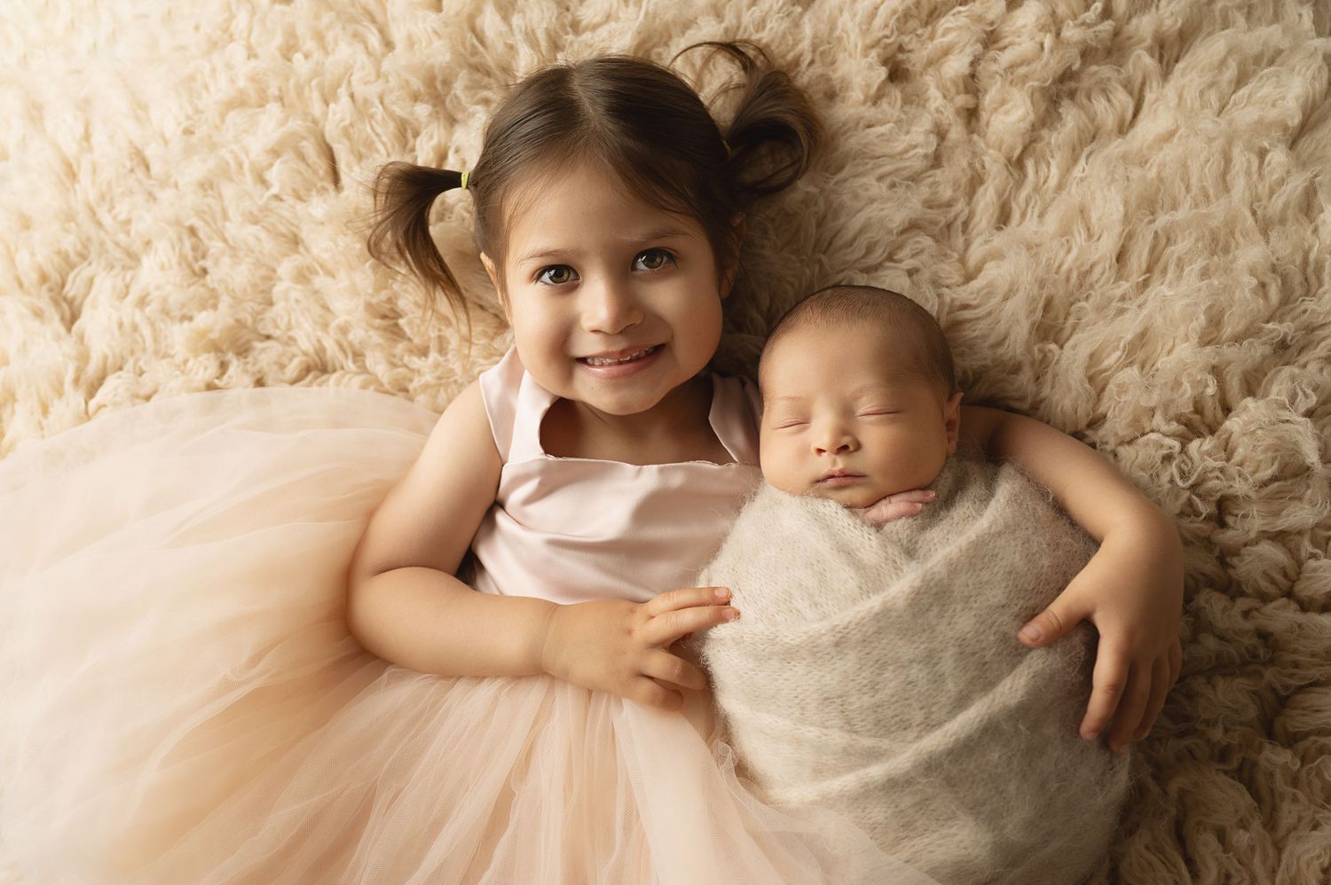 Siblings pose together on cream rug with older sister in tulle dress next to swaddled newborn.