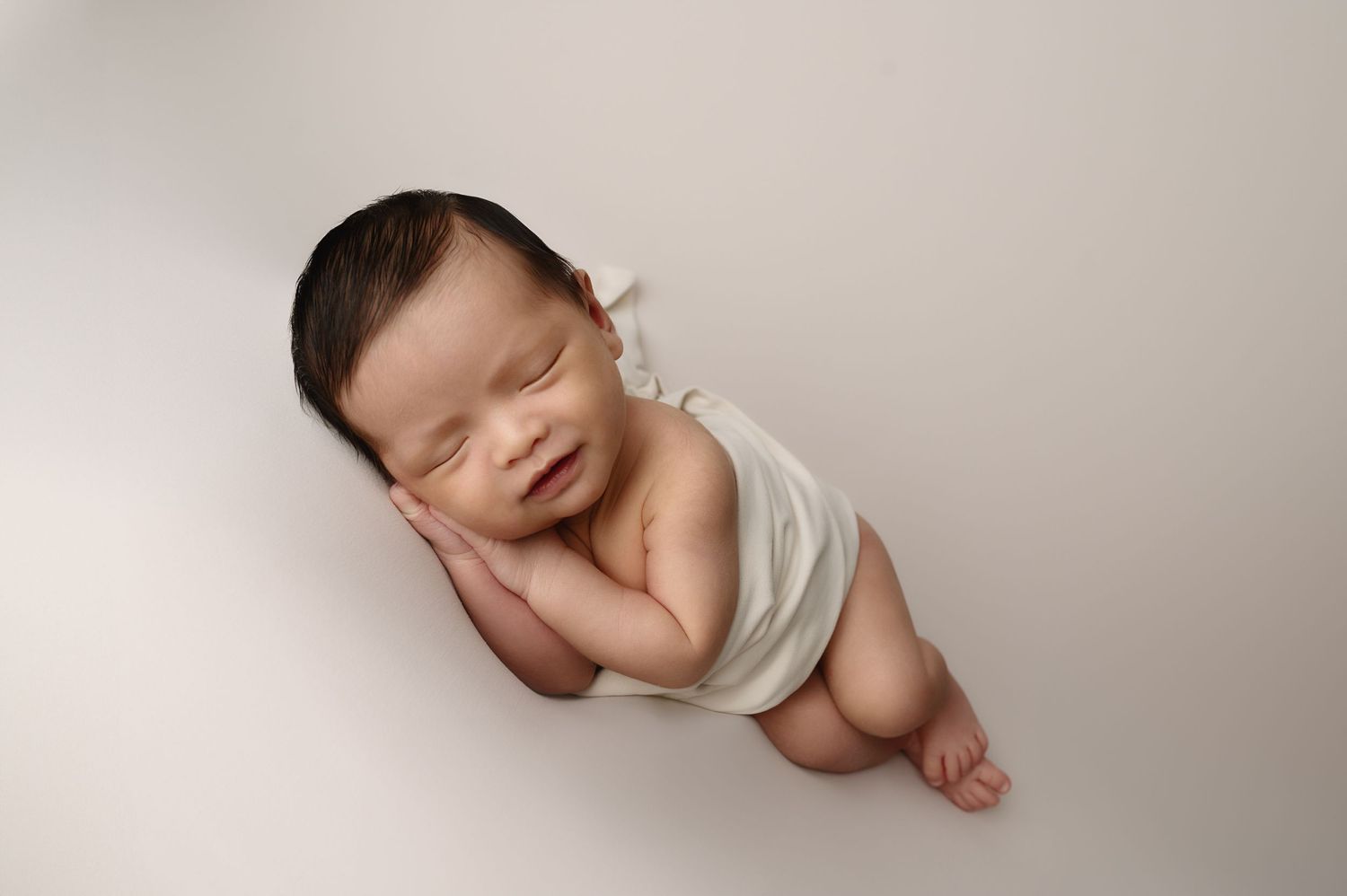 Peaceful newborn partially wrapped in white fabric sleeping on neutral backdrop.