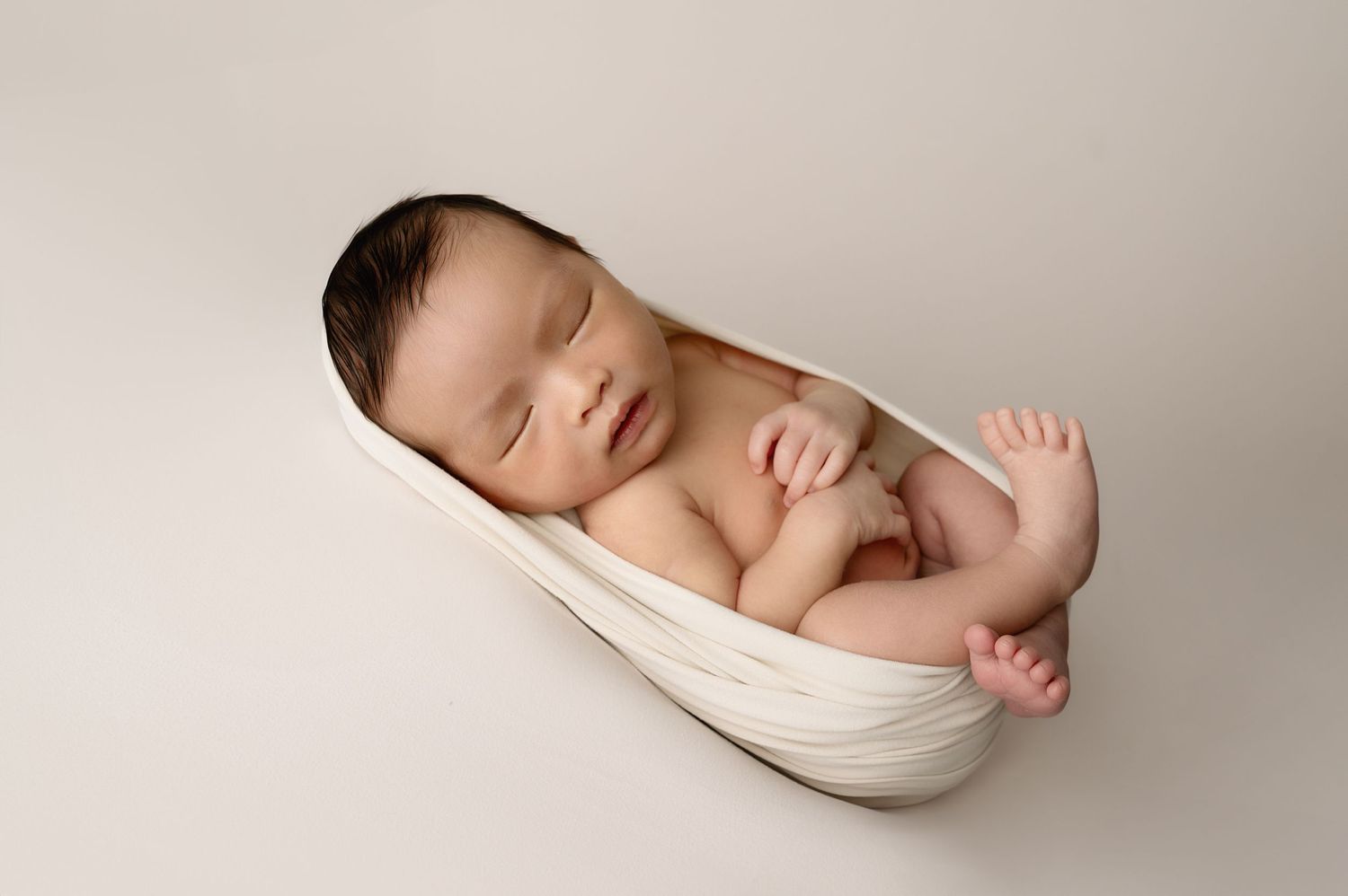 Sleeping newborn posed in cream wrap on neutral backdrop during professional photoshoot.