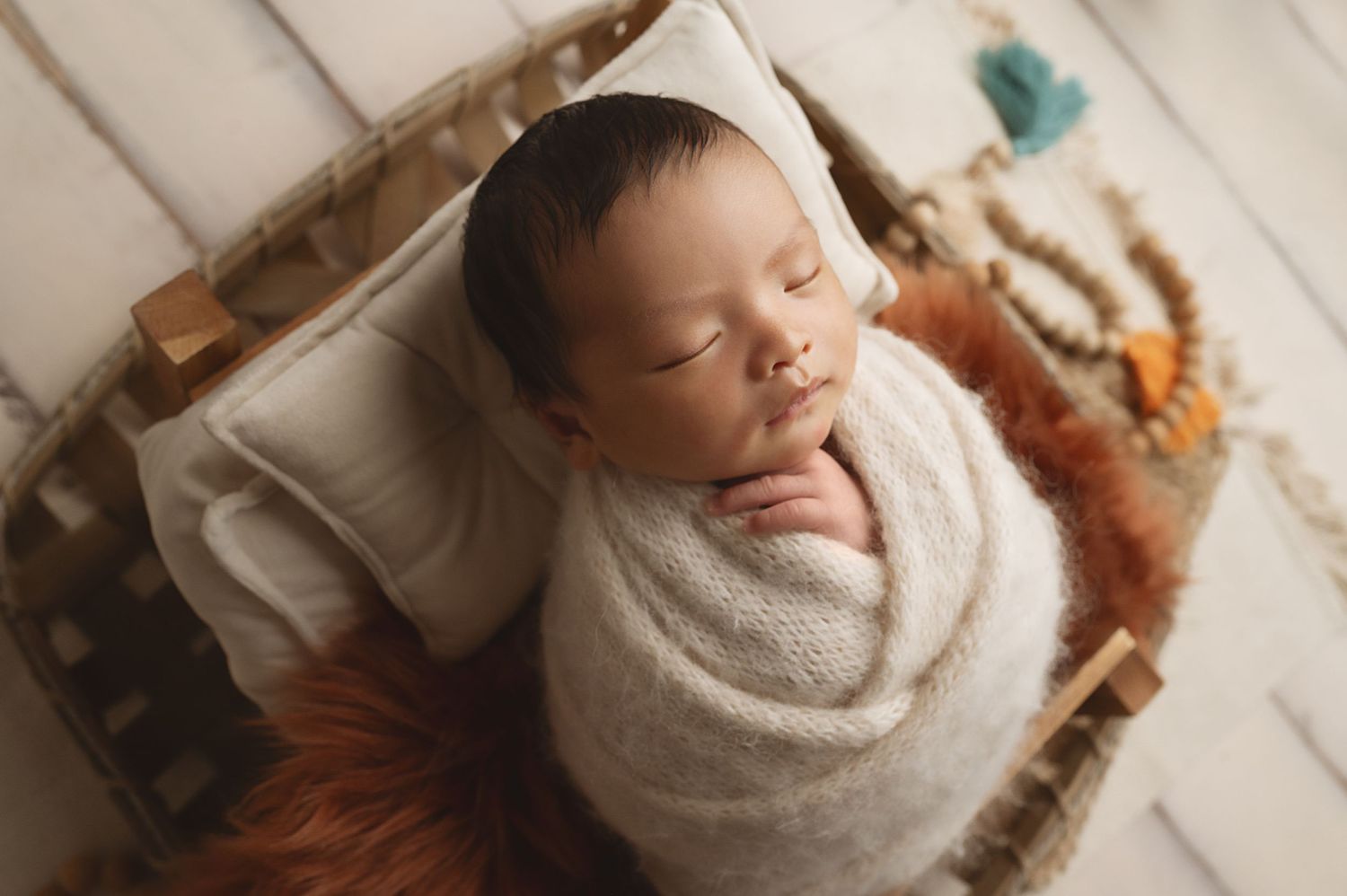 Sleeping newborn wrapped in white swaddle lying in rustic wooden basket with orange fabric.