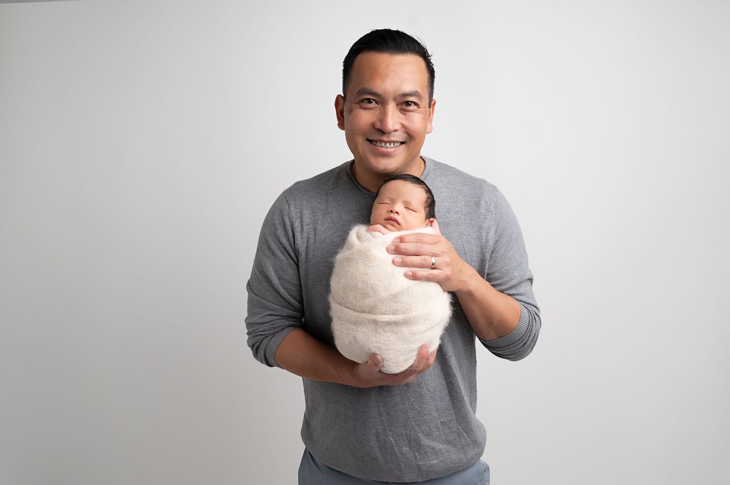Caring parent in grey t-shirt holding swaddled newborn baby against white background.