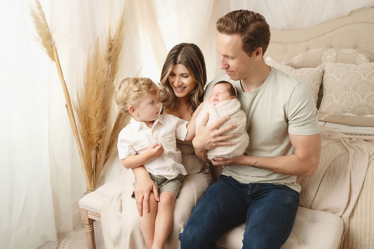 Family portrait with young child and newborn baby sitting together on beige couch with pampas grass decor.