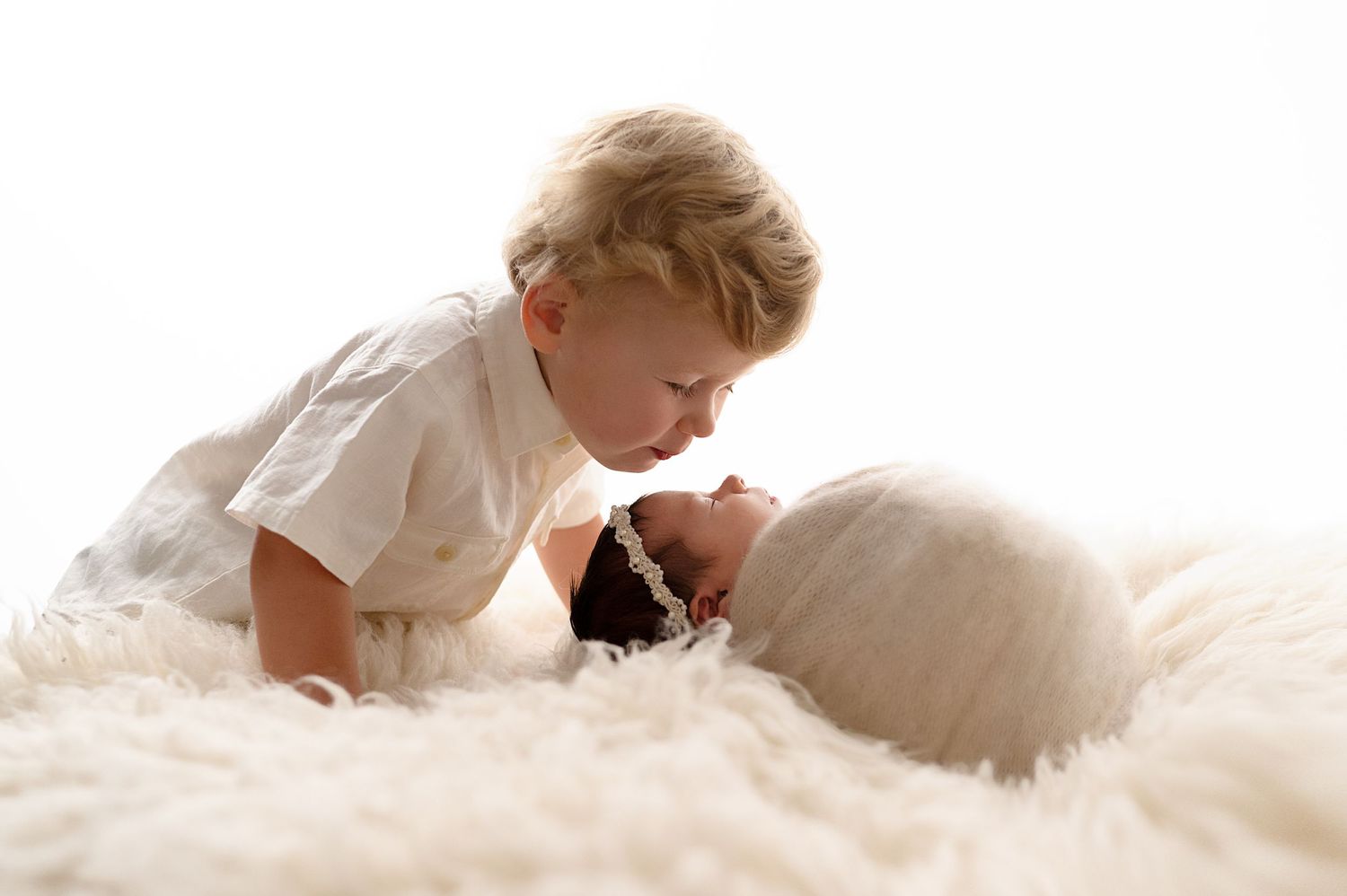 Toddler sibling gently interacting with newborn on soft white furry backdrop.