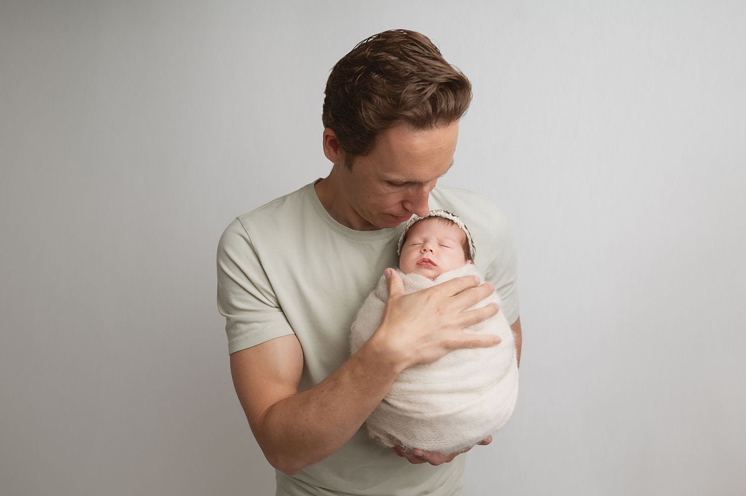 Parent in light grey shirt tenderly holding sleeping newborn baby against neutral wall.