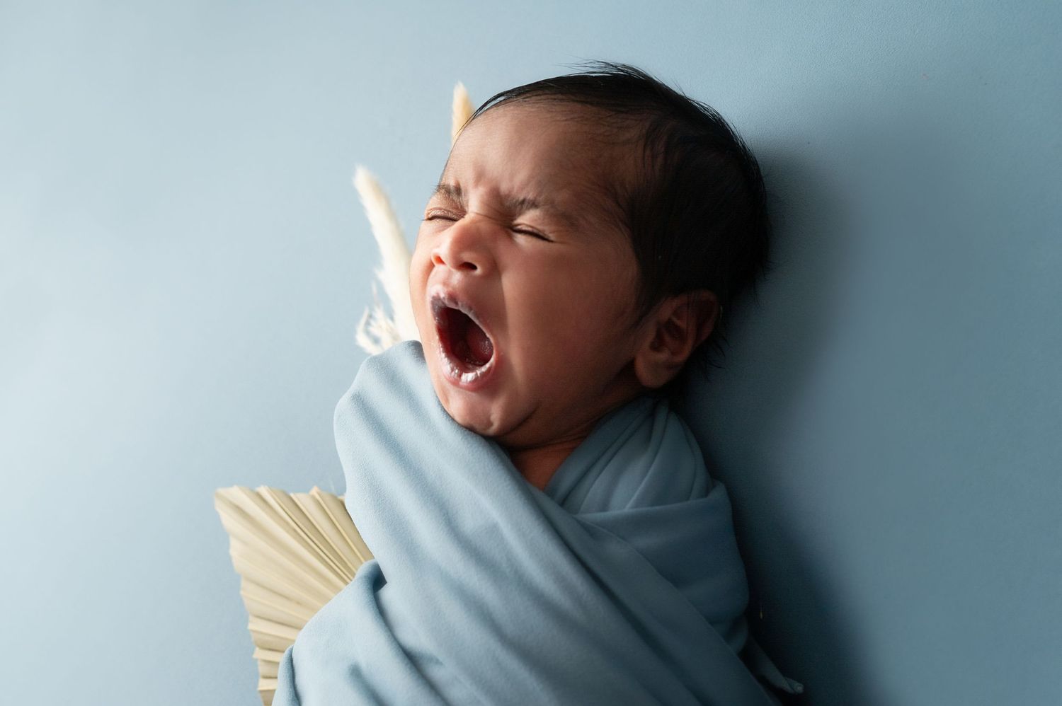 Newborn baby wrapped in blue blanket yawning against light blue background.