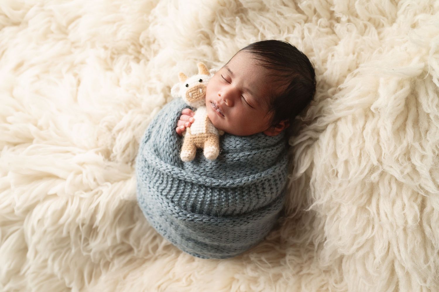 Sleeping newborn wrapped in grey knit blanket with tiny stuffed toy on fur rug.