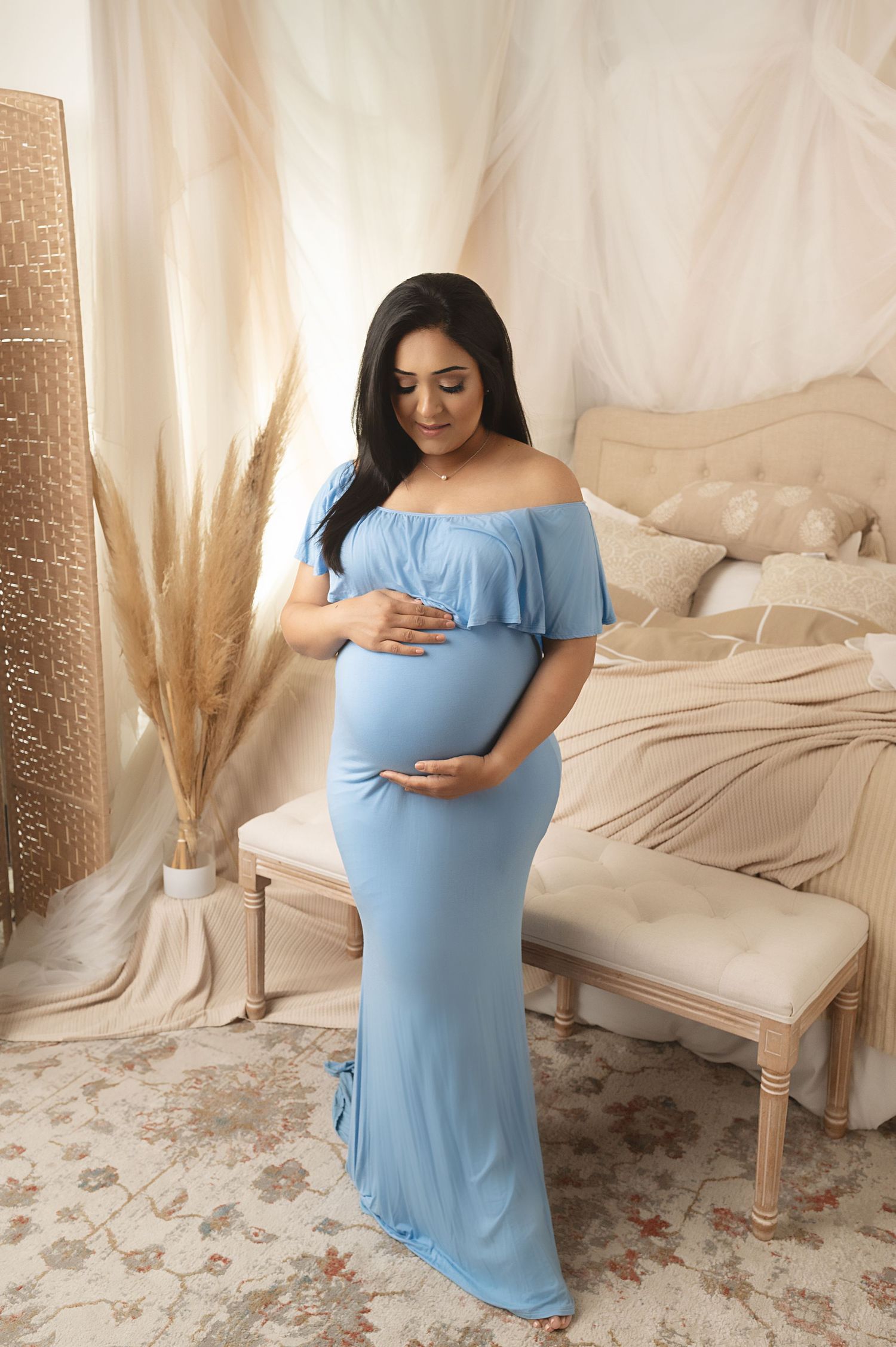 Expectant mother in flowing blue dress poses in front of draped fabric and pampas grass decor.