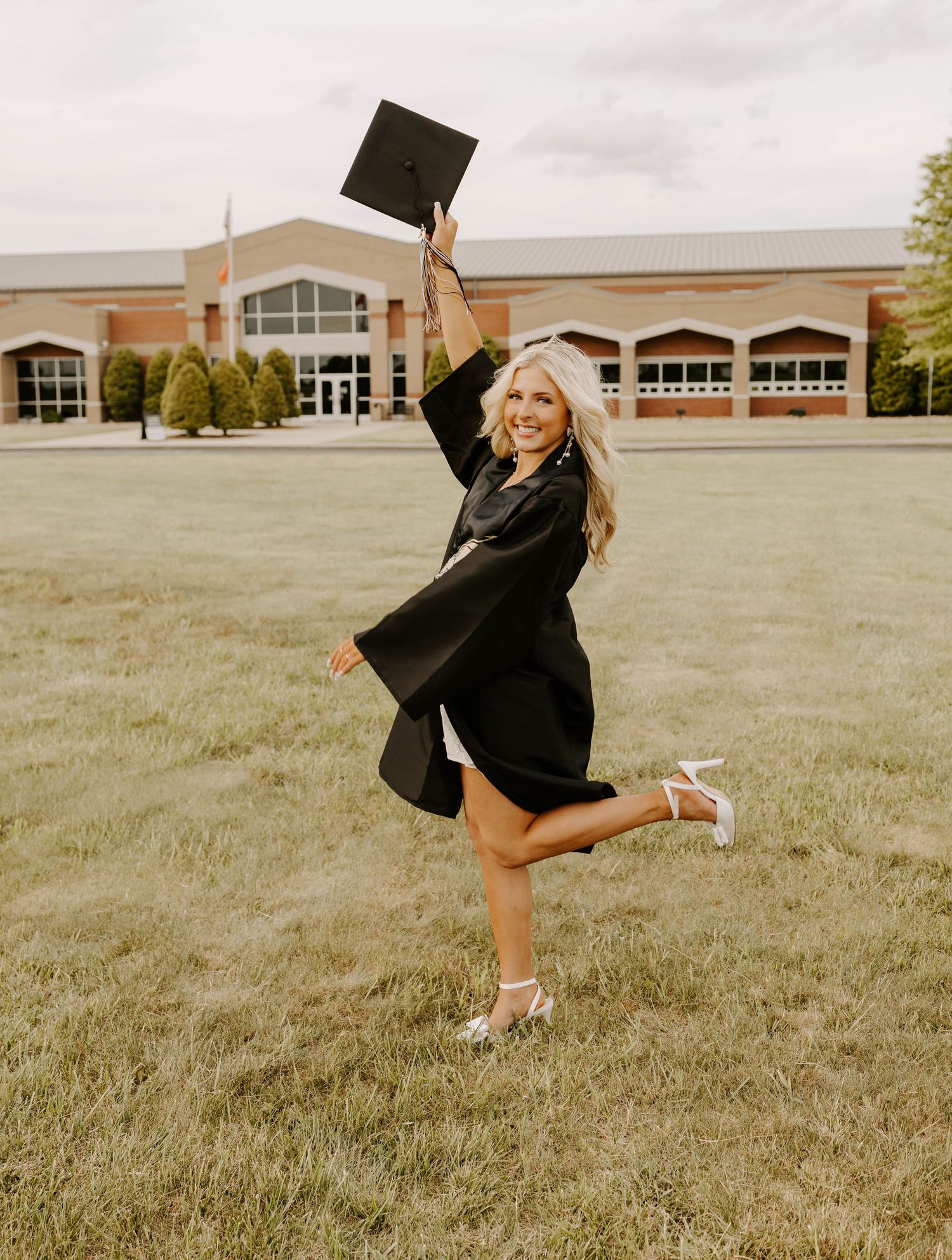 A graduate in black cap and gown jumps joyfully while holding diploma in front of school building.