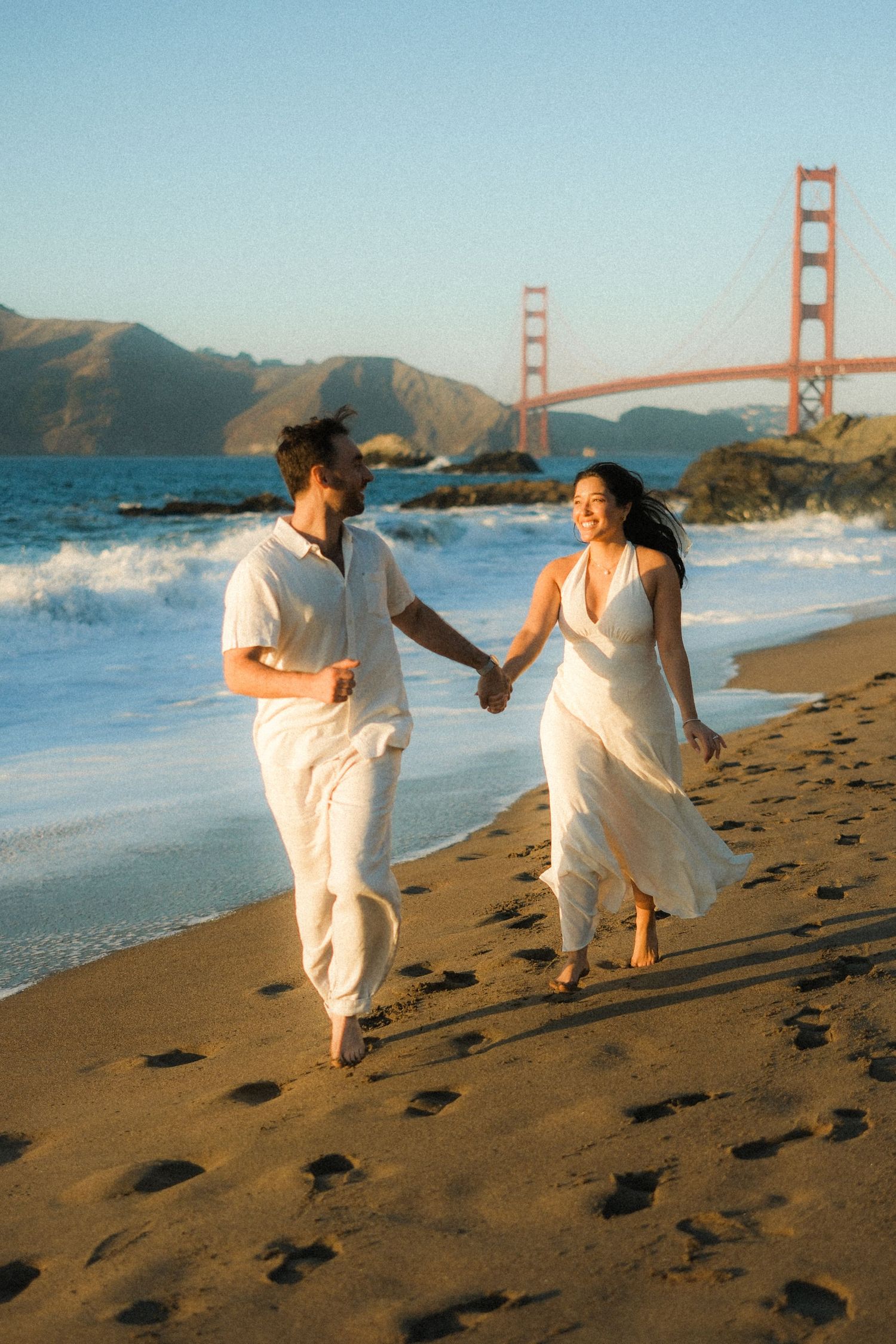 Couple runs hand-in-hand along beach with Golden Gate Bridge in background during sunset.
