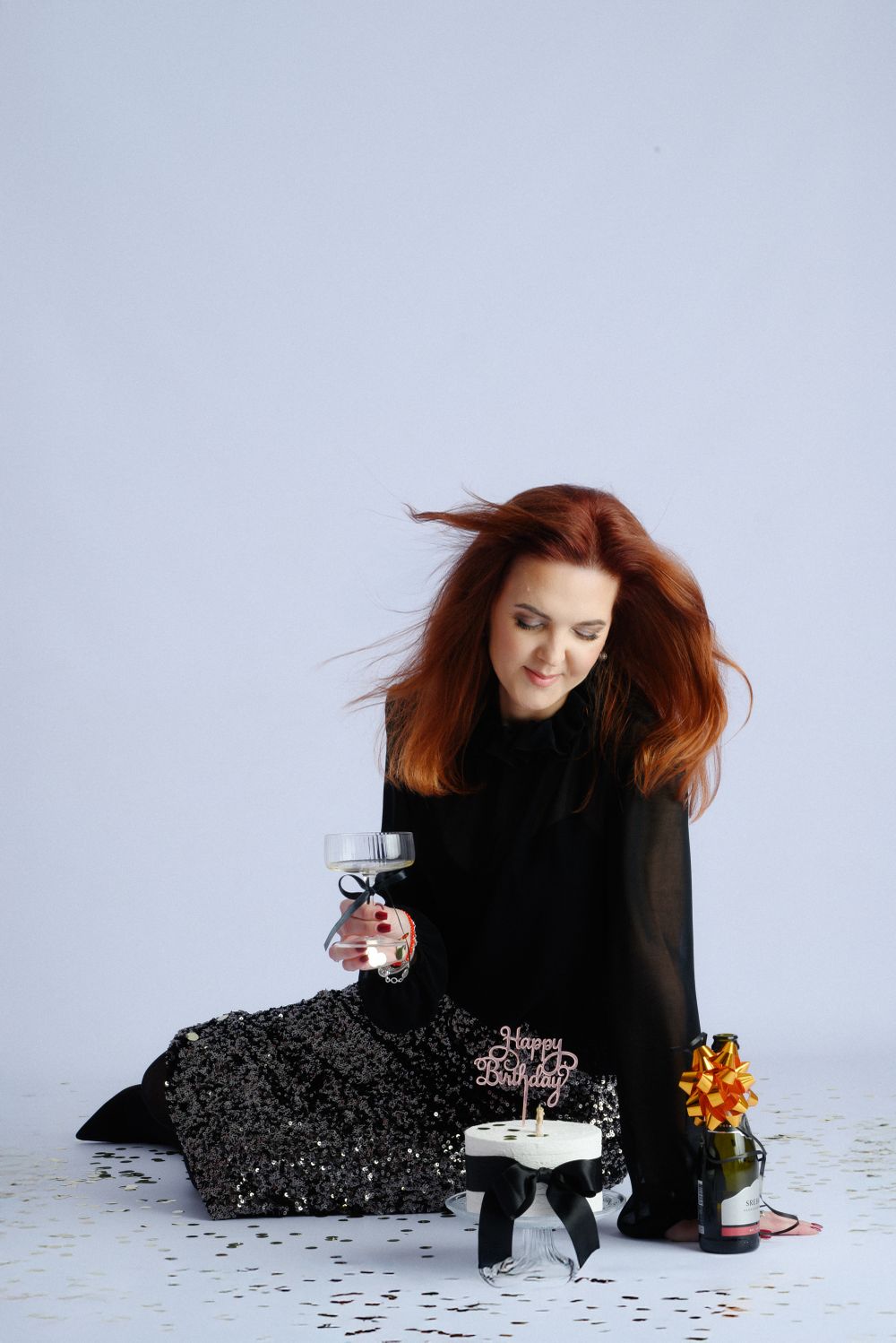 Woman in black dress sitting with wine glass and birthday cake on white floor with confetti.