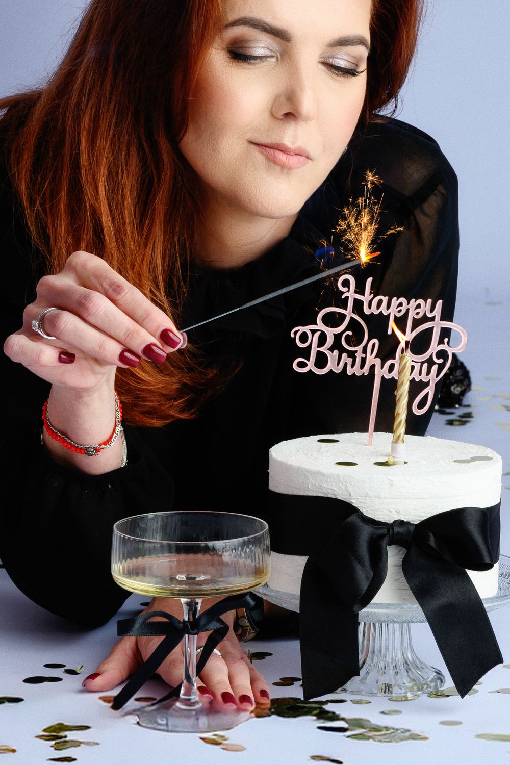 Birthday cake with sparkler and champagne glass on white surface with gold confetti.