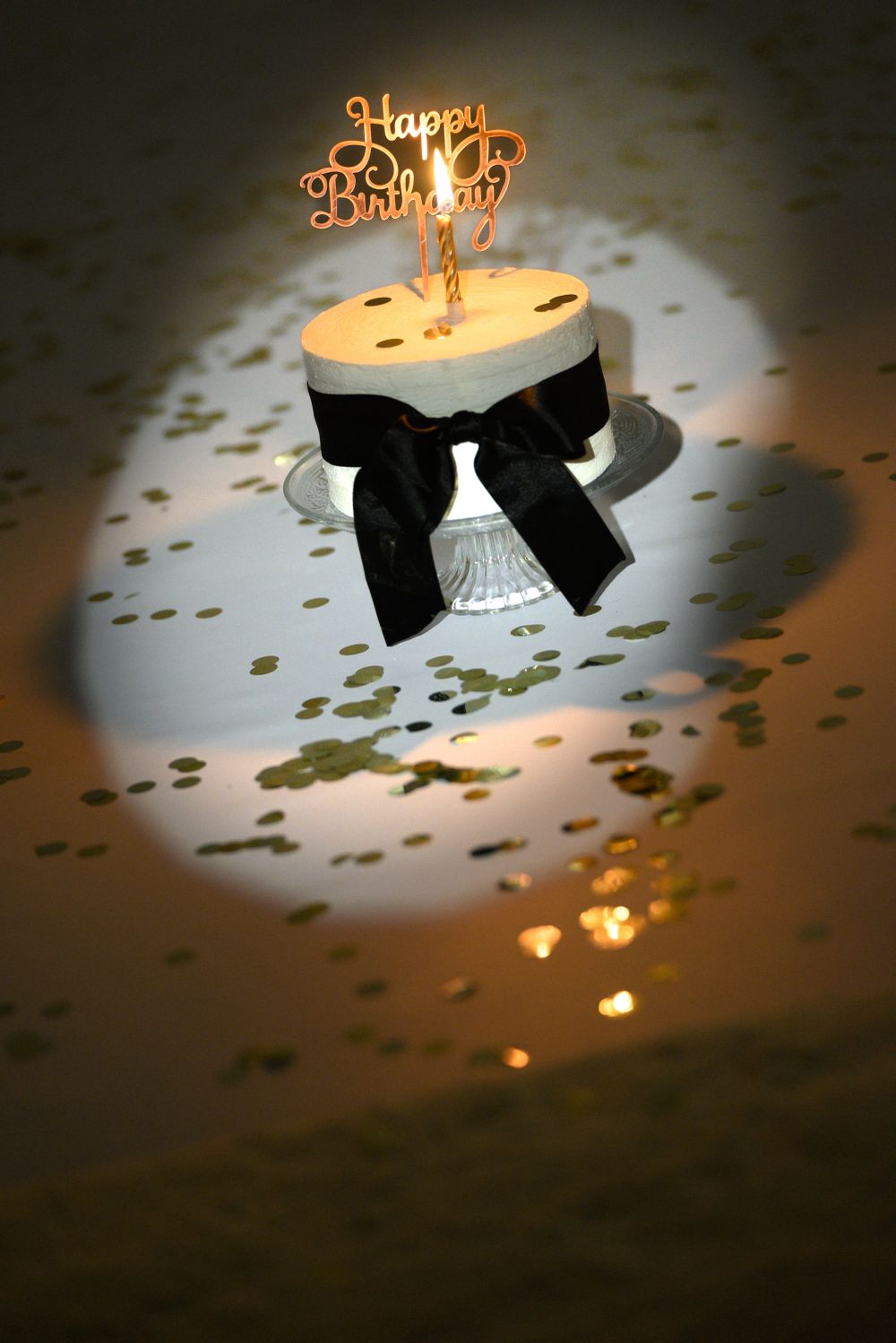 White birthday cake with black bow and glowing happy birthday topper surrounded by gold confetti.