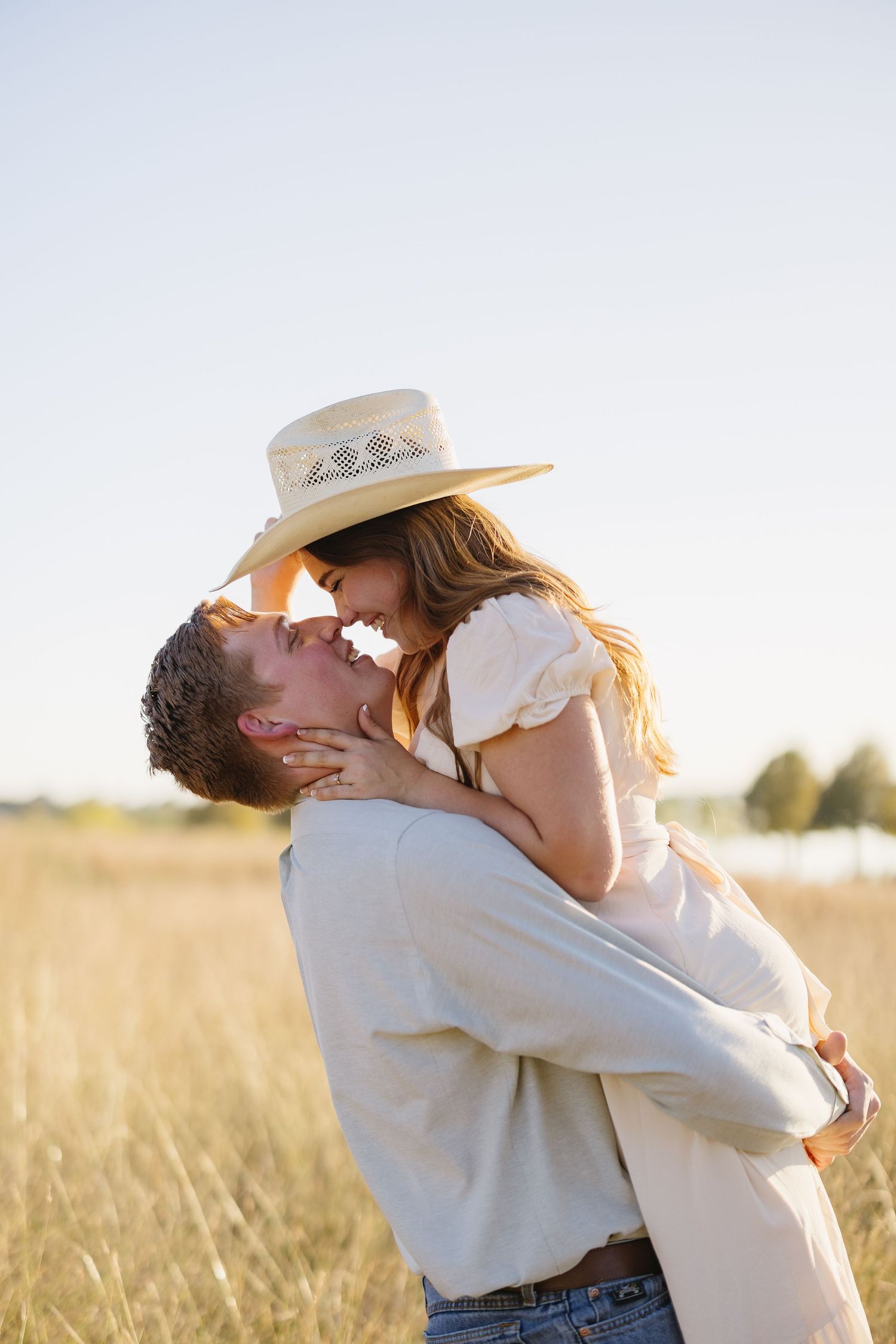 Man lifting woman in straw hat for a kiss in a golden sunlit open field during sunset.