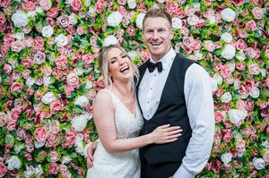 A happy newlywed couple poses together in front of a beautiful floral wall backdrop of pink and white roses.