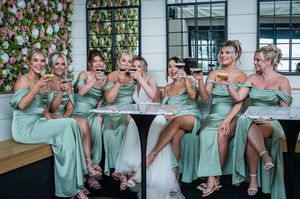 A group of bridesmaids in matching mint green satin dresses share laughter while seated at a restaurant table.