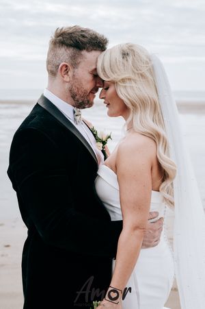 A romantic wedding couple shares an intimate moment on a cloudy beach, with the bride in white and groom in black.