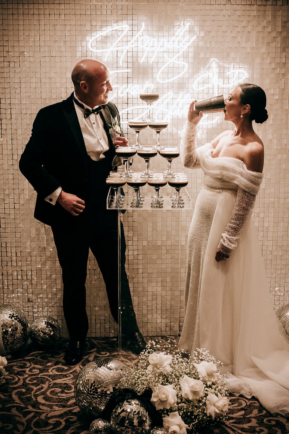 Bride and groom toast champagne tower at elegant wedding reception with neon sign and disco ball decor.