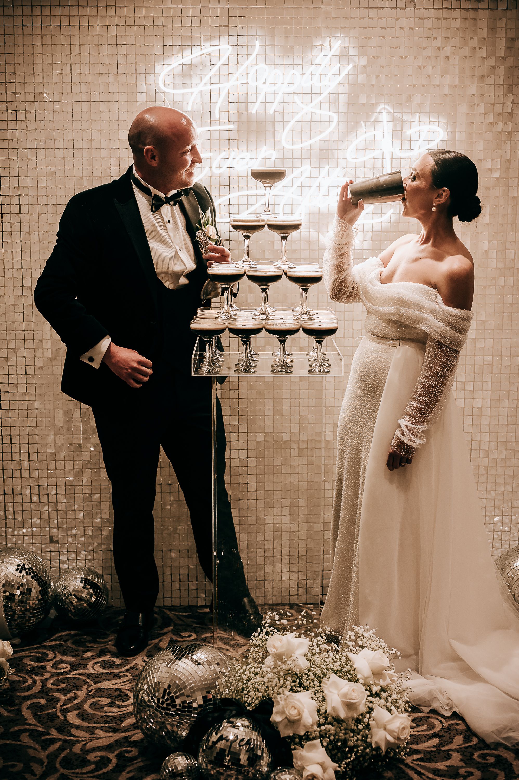 Bride and groom toast champagne tower at elegant wedding reception with neon sign and disco ball decor.
