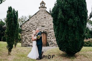 A couple poses in front of a rustic stone church with cypress trees creating an intimate outdoor wedding scene.