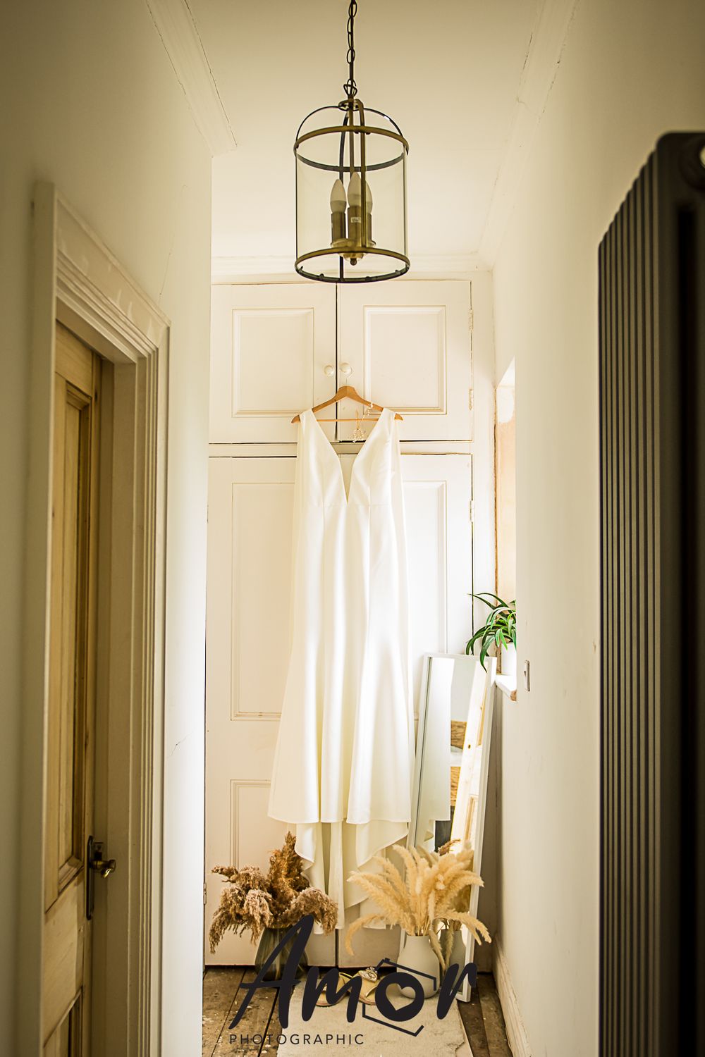 A white wedding dress hangs elegantly beneath a vintage lantern in a bright hallway.