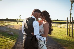 A romantic wedding couple shares a tender moment at sunset on a rural path lined with wooden fences and trees.