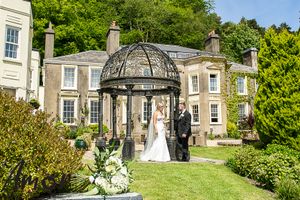 A couple stands under an elegant domed gazebo in front of a grand stone manor house surrounded by manicured gardens.