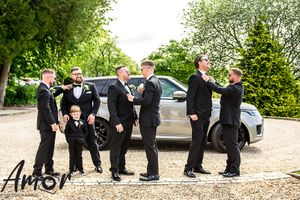 Groomsmen in black tuxedos pose playfully in a line outdoors with greenery and a car in the background.