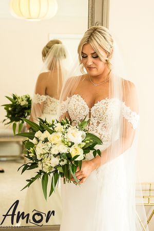 Elegant bride in lace white gown holds romantic white and green bouquet while looking down during wedding preparation.