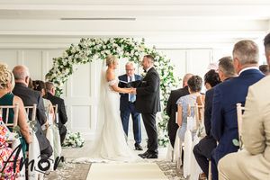 Wedding ceremony taking place under a white floral arch with guests seated in chairs at an elegant indoor venue.