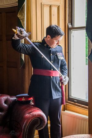 Military officer in formal dark uniform with red sash adjusts ceremonial sword while standing by window and leather chair.