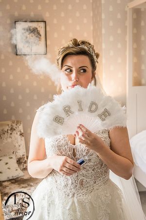 A bride in a white lace wedding dress holds a delicate feathered fan in front of her face in a vintage-style setting.