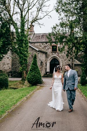 Romantic couple walks along stone church pathway surrounded by lush greenery in elegant wedding attire.