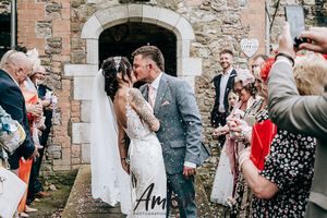 A wedding celebration outside a stone church with guests throwing confetti as the newlyweds share a kiss.