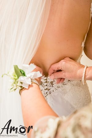 Close-up of hands adjusting a bridal dress detail with a white rose and greenery corsage in view.