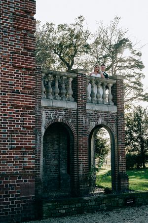 An ornate brick archway with stone balustrades stands in a garden setting with trees visible through the opening.