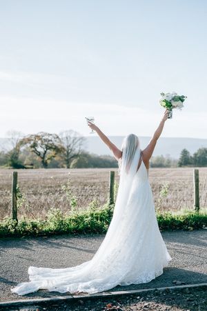 A flowing white wedding dress train spreads gracefully across a country path as someone poses with arms raised against scenic hills.