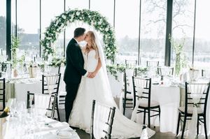 Indoor wedding reception venue with elegant floral arch, white chairs and waterfront view through floor-to-ceiling windows.