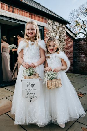 Two flower girls wearing white lace dresses and fur shawls hold baskets with flowers for a winter wedding ceremony.