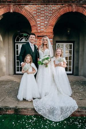 Wedding party wearing white dresses poses together against charming brick building with arched doorways and windows.