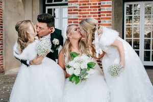Three brides in white wedding dresses pose together while holding bouquets on the porch of a brick building.