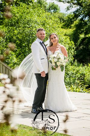 A bride and groom on their wedding day pose together outdoors on a stone patio surrounded by lush greenery.