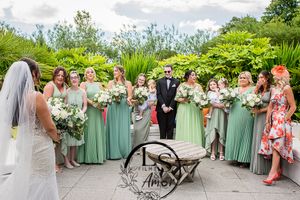 A wedding party wearing sage green dresses poses outdoors in a garden setting holding white floral bouquets.