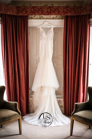A white lace wedding dress hangs elegantly between red curtains with two velvet chairs positioned symmetrically.